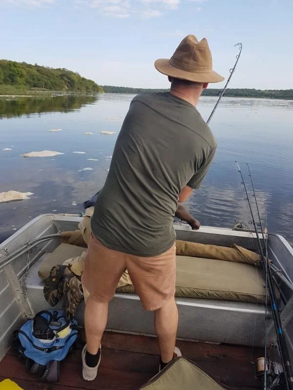 A man in a green T-shirt, tan shorts, and a wide-brimmed hat stands on a boat, fishing in a calm lake with trees in the background.