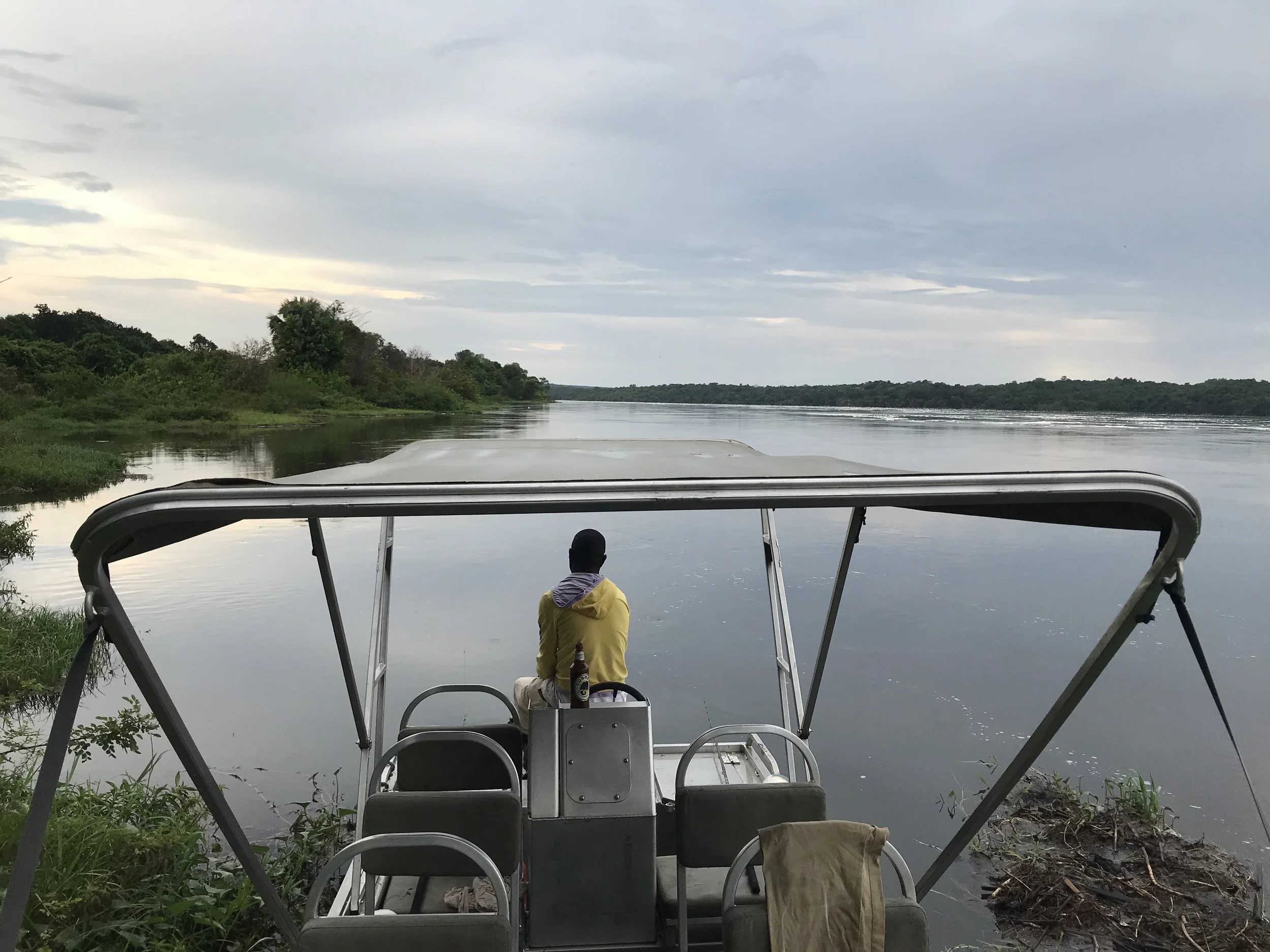 View from a boat on a river, with a man sitting at the back, looking out at the water and the landscape. The sky is overcast with clouds, and there is lush green foliage along the riverbanks.