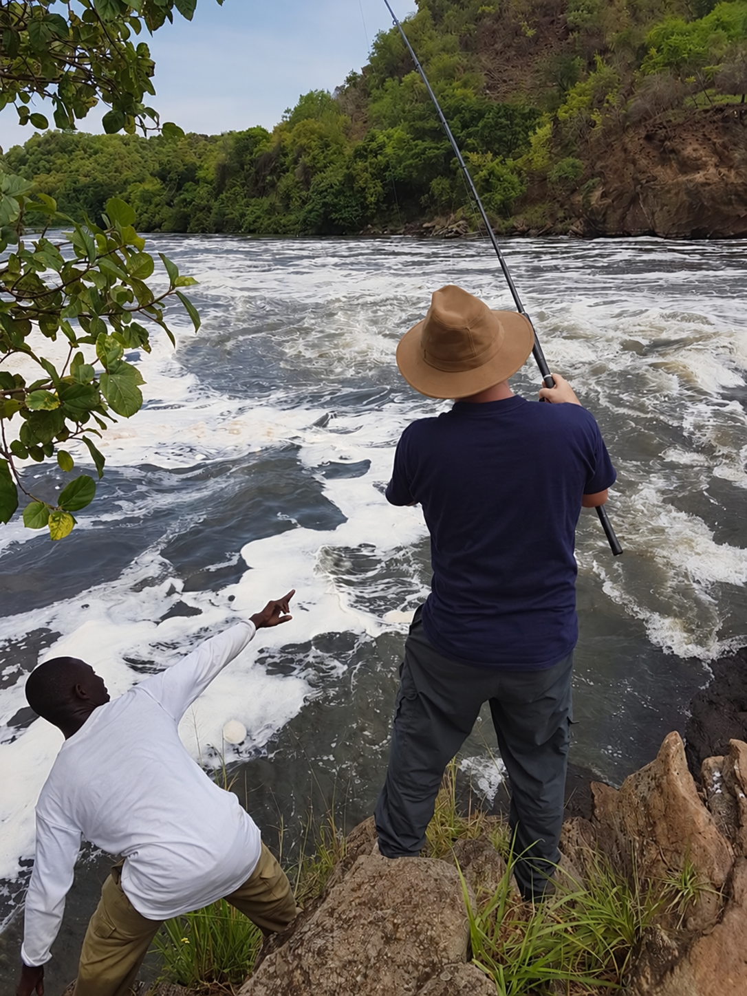 Two men by a river, one fishing with a rod, the other pointing towards the water, surrounded by green trees and rocky terrain.