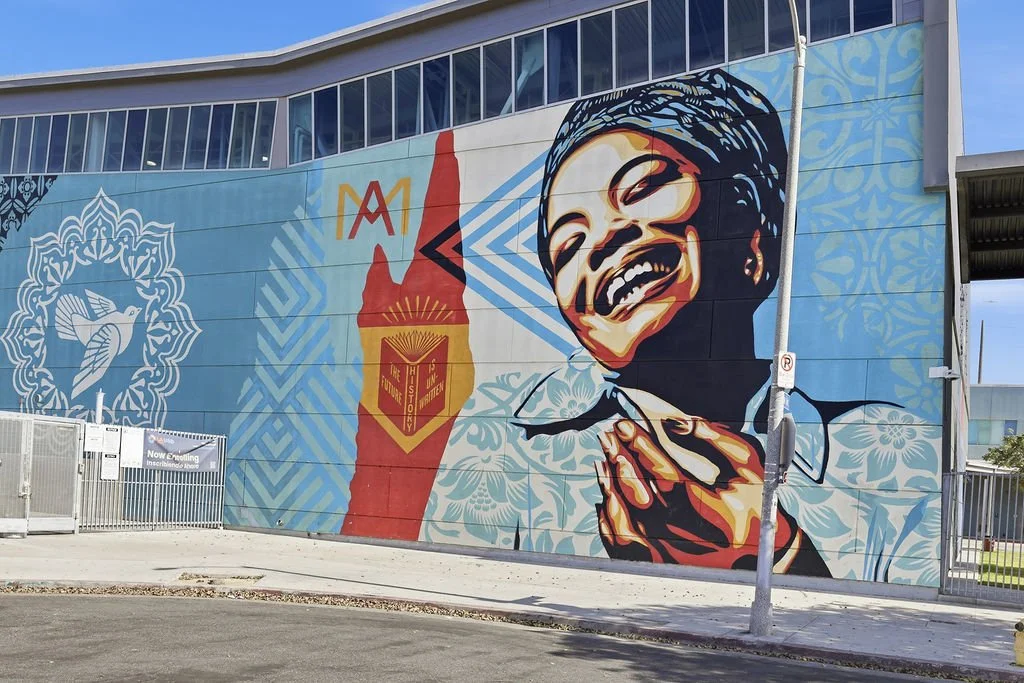 A colorful mural on a building wall featuring a laughing woman holding a spoon, with abstract patterns, a stylized flame, a bird, and the text 'The Future is Written' on a red background.