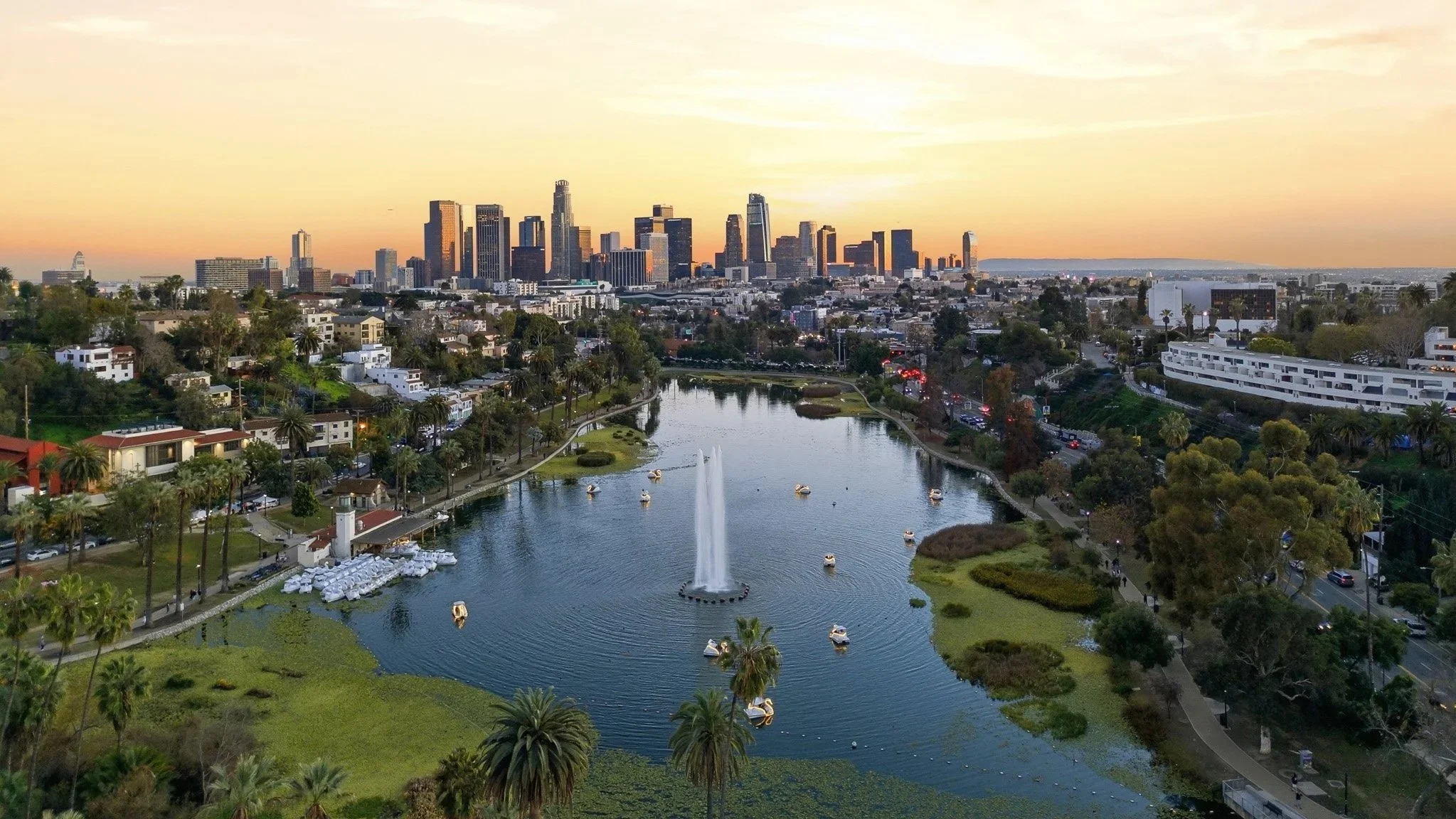 A city skyline at sunset with tall buildings, a waterway with boats, and lush green trees in the foreground.