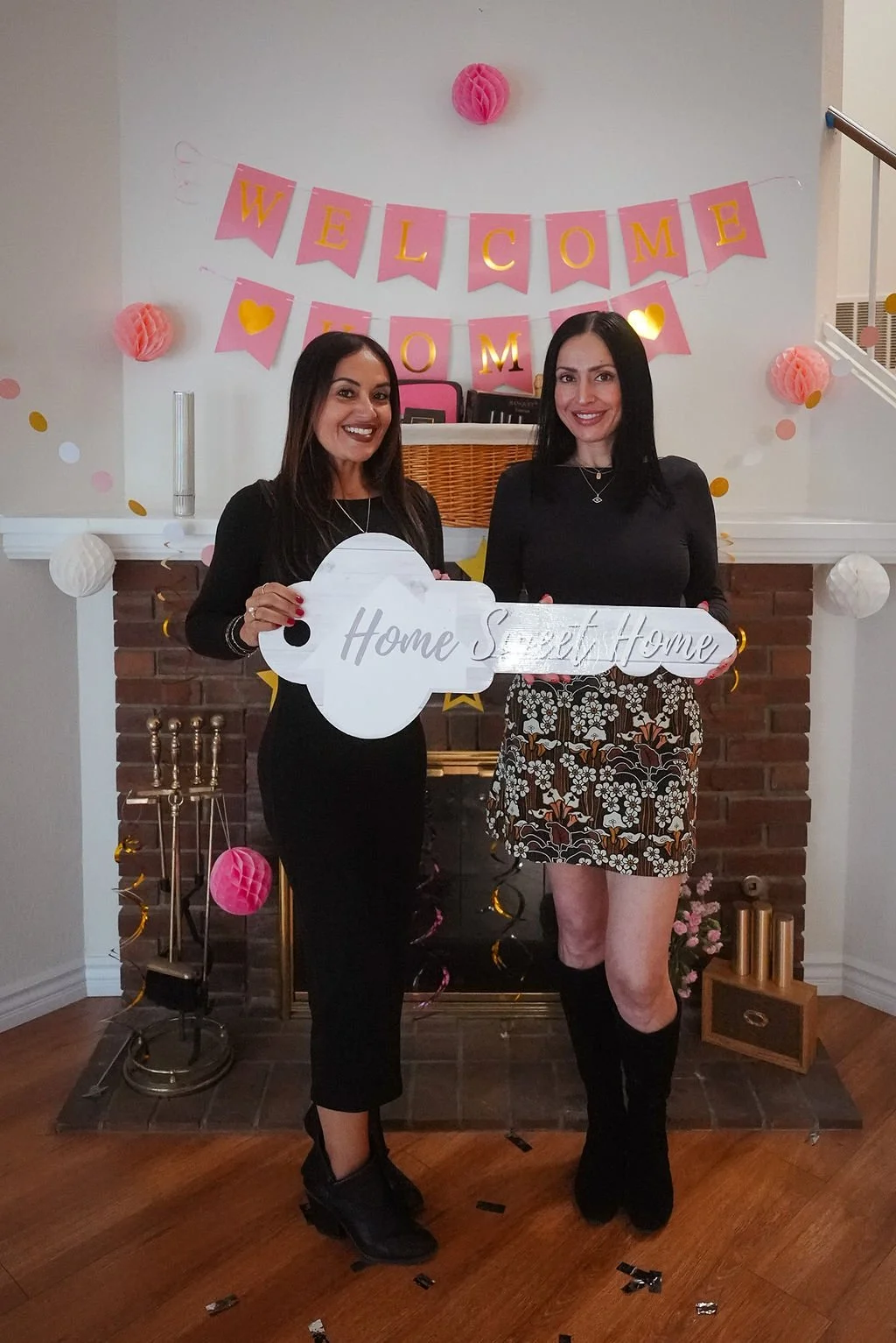 Two women standing in front of a decorated fireplace with a pink and gold 'Welcome Home' banner, holding a sign that says 'Home Sweet Home', celebrating a homecoming event.
