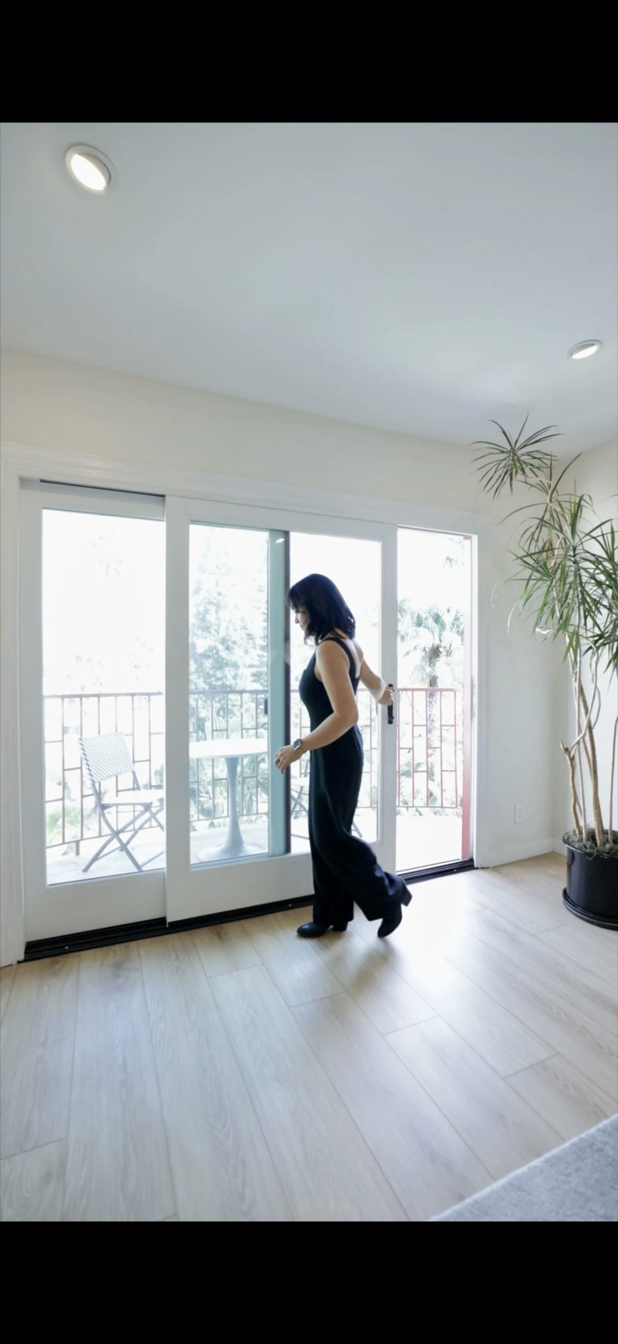 A woman in black clothes opening a sliding glass door to a balcony with outdoor chairs and a small table.