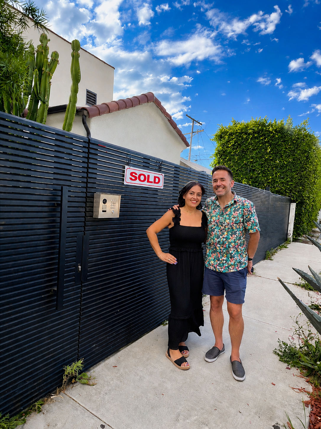 A smiling couple standing outside a black gate with a "SOLD" sign, on a sunny day with a bright blue sky and white clouds, in front of a white building with a red-tiled roof and some green plants.