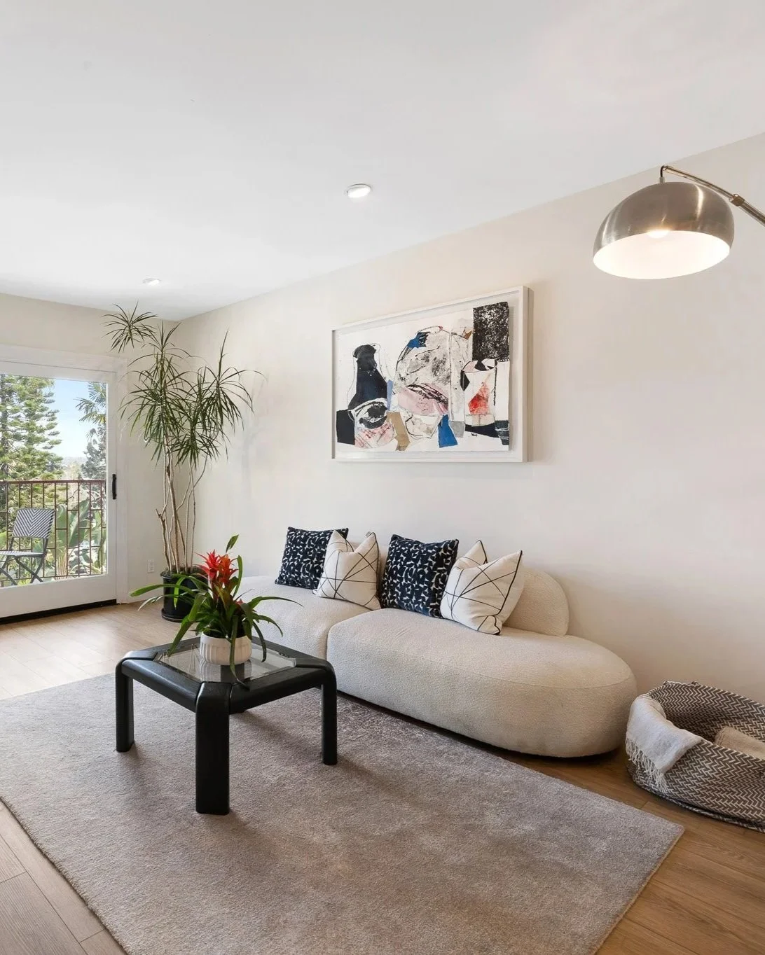 Living room with white curved sofa, black coffee table, large floor plant, abstract wall art, and window with outdoor view