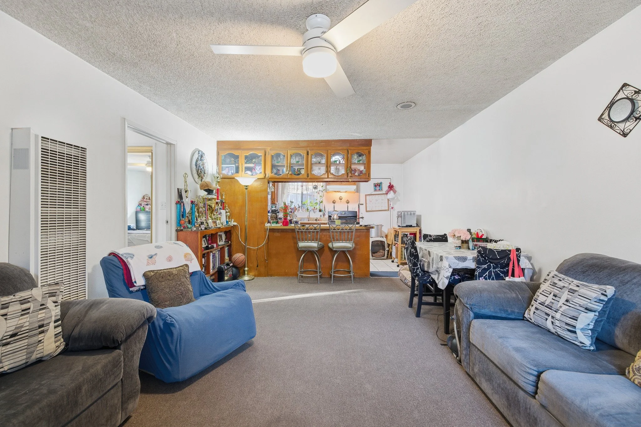 A cozy living room with gray sofas, a blue armchair, and a dining table with chairs, decorated with pillows and floral arrangements, with a view into the kitchen in the background.