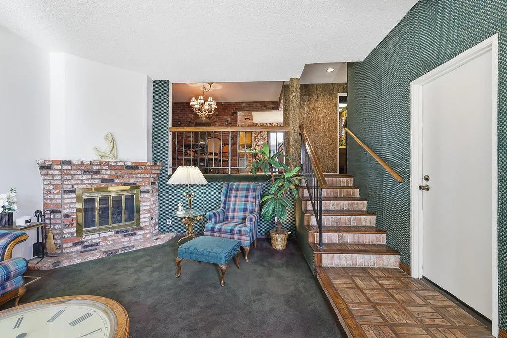 Living room with fireplace, plaid armchair, side table, floor lamp, potted plant, staircase, and green patterned walls