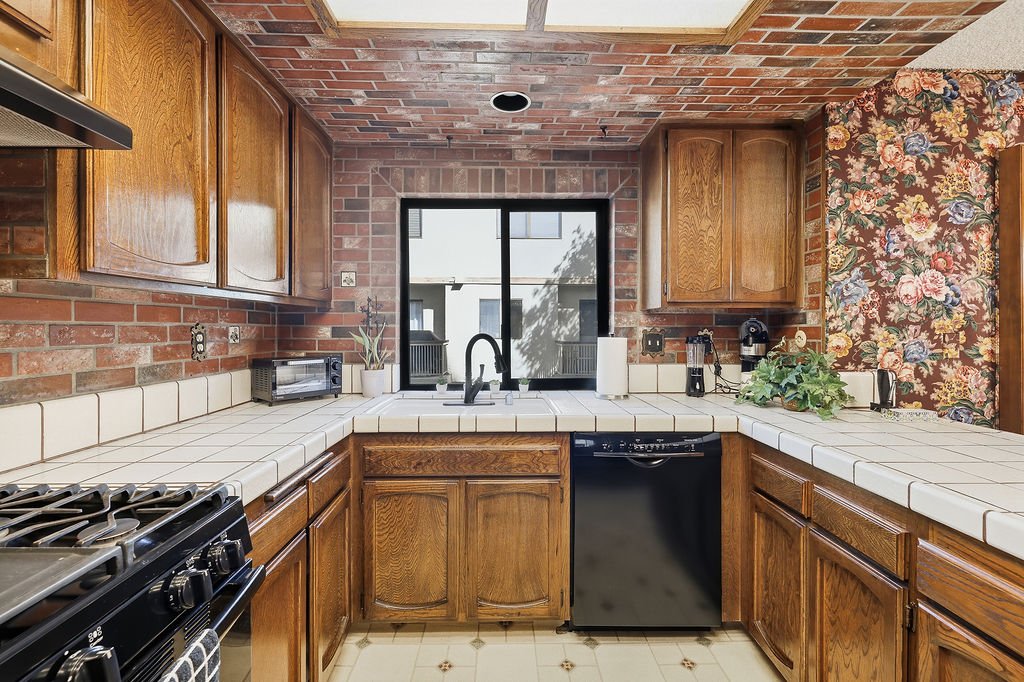 Kitchen with wooden cabinets, white tiled countertops, a black dishwasher, and a brick wall background, with a window above the sink.