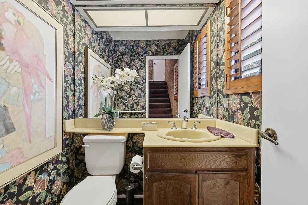 Small bathroom with dark floral wallpaper, wooden window shutters, cream sink, and a toilet with a flower vase on the shelf above.