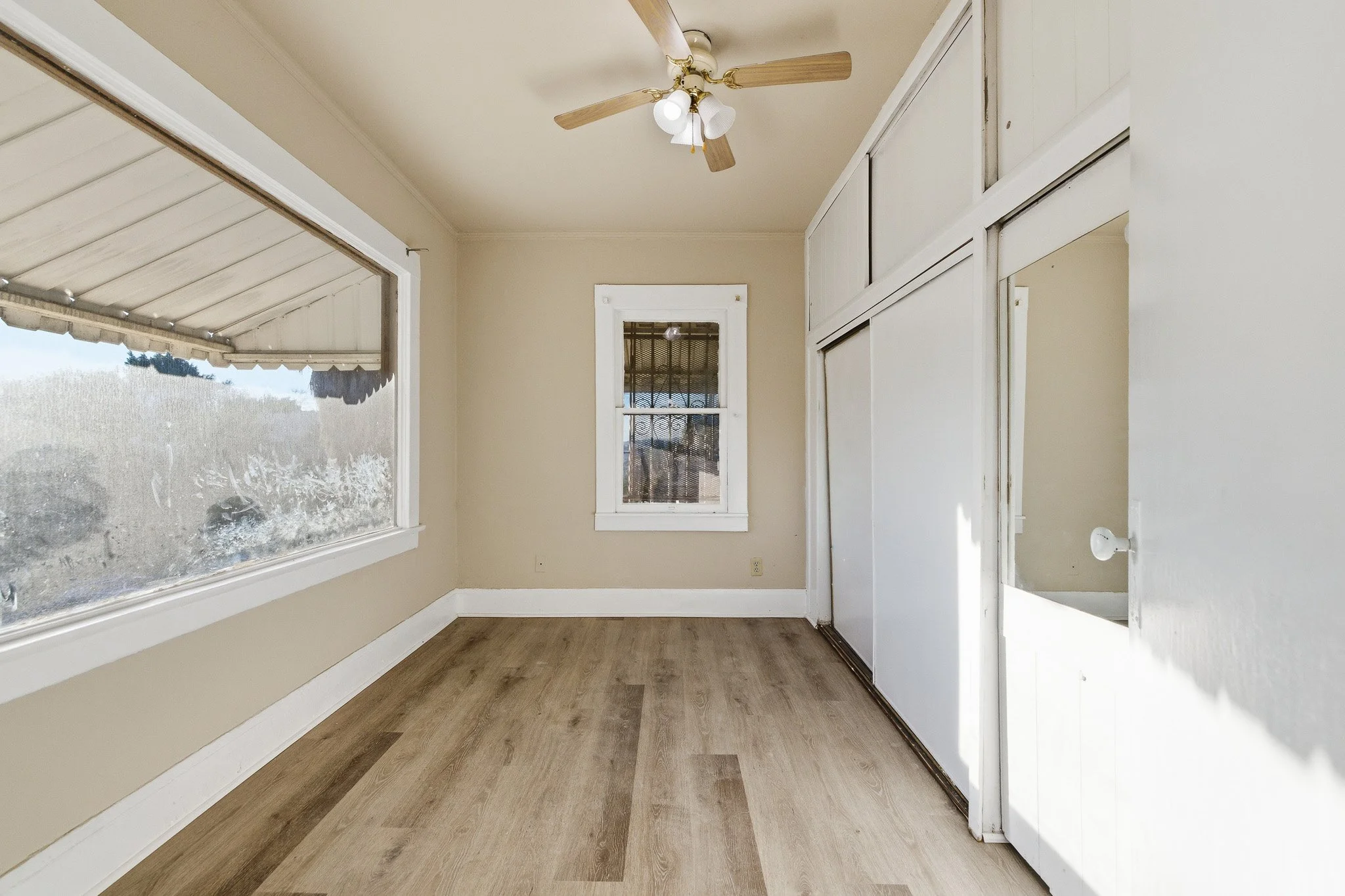 Empty room with beige walls, wooden floor, two windows with natural light, ceiling fan with four blades, and white sliding closet doors.