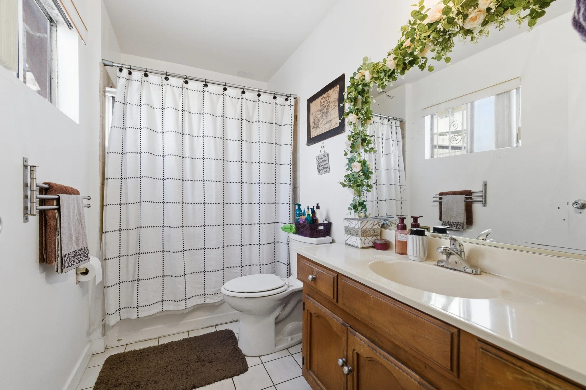 A bathroom with white walls, a wooden vanity with a white countertop, a mirror above it, two small windows, a shower with a white and black grid curtain, a toilet, a brown rug, towels on a wall rack, and a decorative ceiling with a floral garland.
