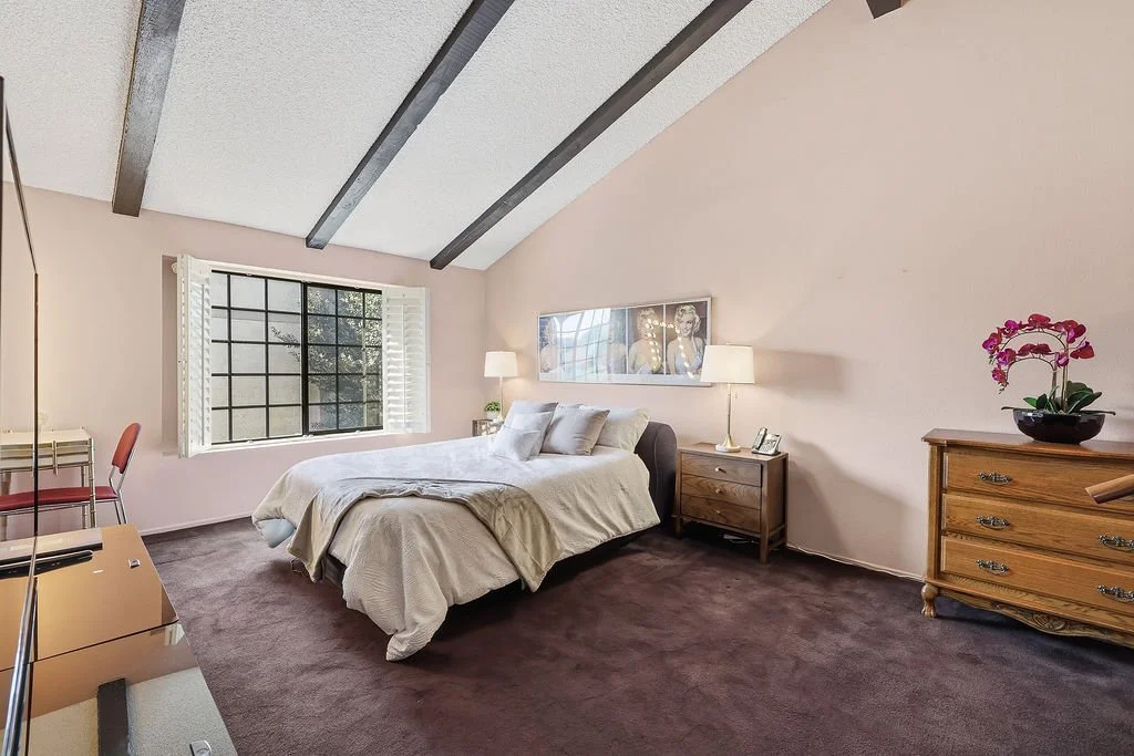 A bedroom with a sloped ceiling featuring wooden beams, a large window with white shutters, a bed with white bedding, bedside lamps, a wooden dresser with a pink orchid, and a small desk with chairs.