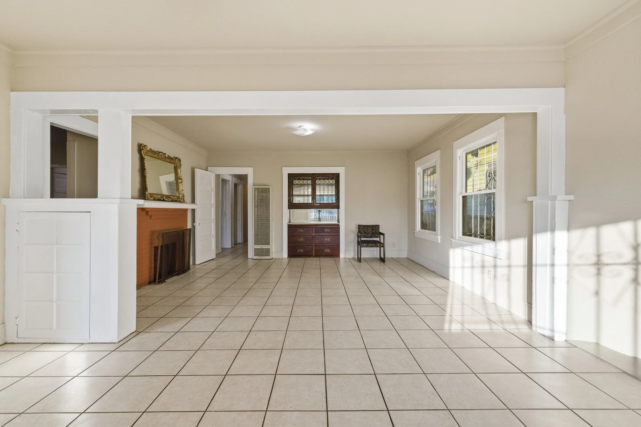 Empty living room with beige walls, tiled floor, and multiple windows letting in natural light. There is a mirror on the fireplace mantel, a small chair, and a chest of drawers.