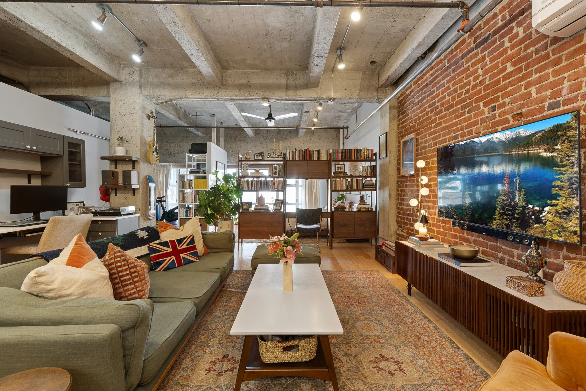 Living room with brick wall, flat-screen TV, bookshelf, ceiling fan, concrete ceiling, various decorations, plants, sofas, and a coffee table.