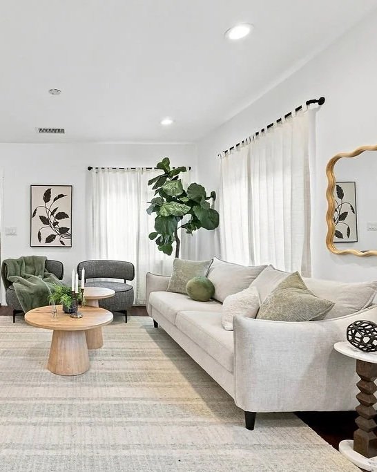 Bright living room with white walls, beige sofa with neutral cushions, large green potted plant, round wooden coffee table, green and gray armchairs, framed botanical art on walls, curtains over windows, and a mirror on the wall.