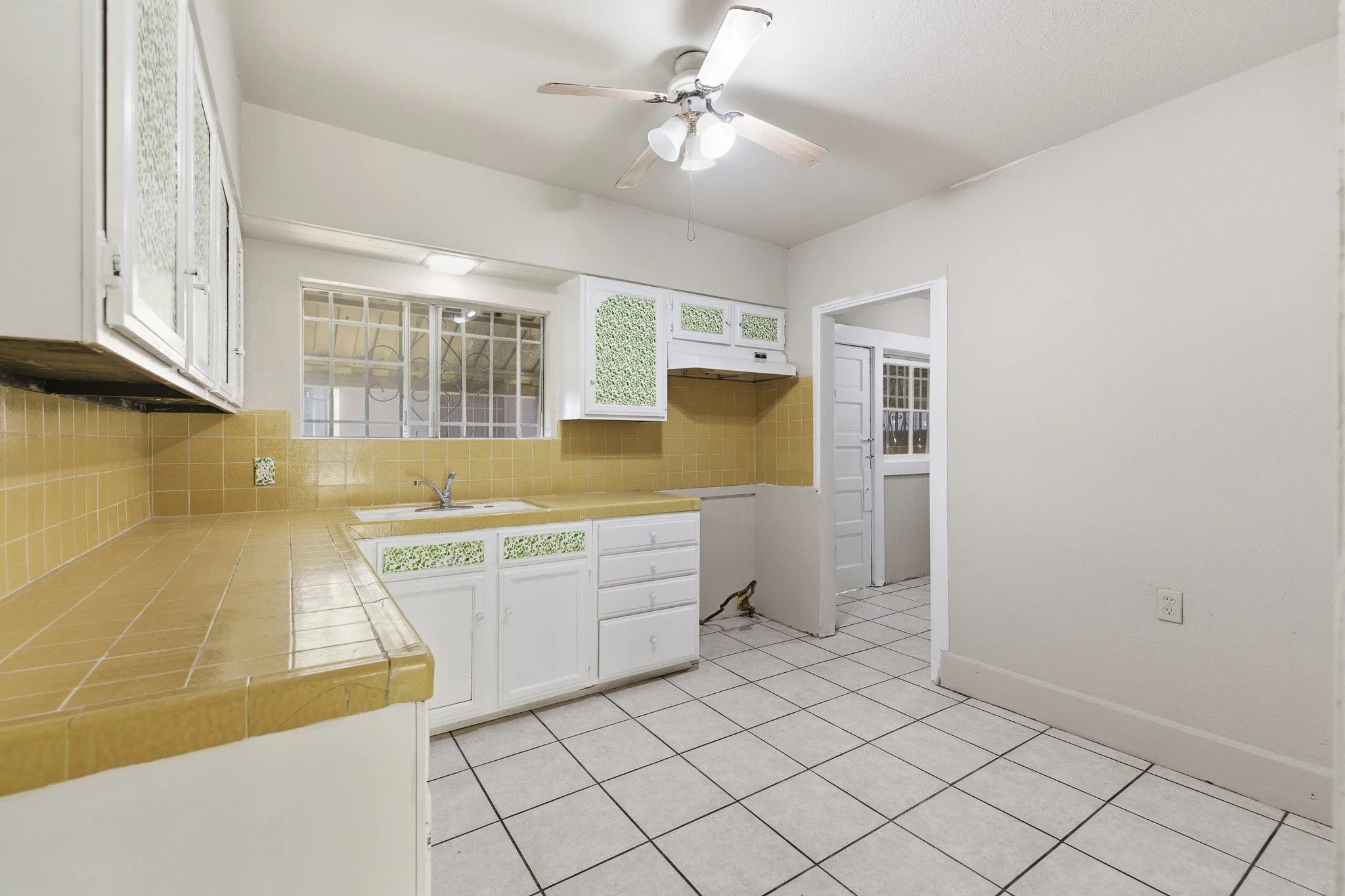 Empty kitchen with yellow countertops, white cabinets with green decorative cutouts, yellow tile backsplash, white tiled floor, and ceiling fan with light fixtures. Doorway leads to another room.