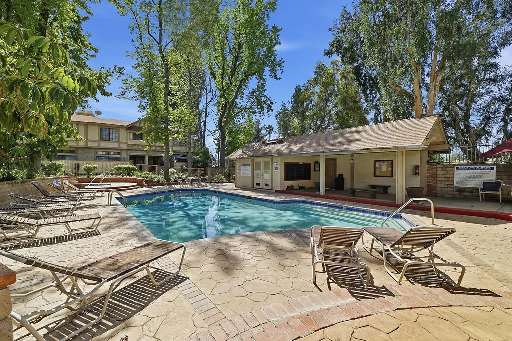 A small outdoor swimming pool surrounded by lounge chairs, trees, and a shaded building with benches, set in a sunny area.