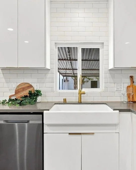 White kitchen sink with gold faucet and handles, surrounded by white cabinets, white subway tile backsplash, window above, leaf and cutting board on counter.