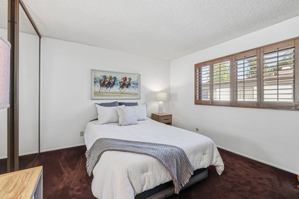 A bedroom with a white bed, gray and white pillows, a nightstand with a lamp, a framed horse artwork, large windows with wooden shutters, and a mirrored closet door, with brown carpet flooring.
