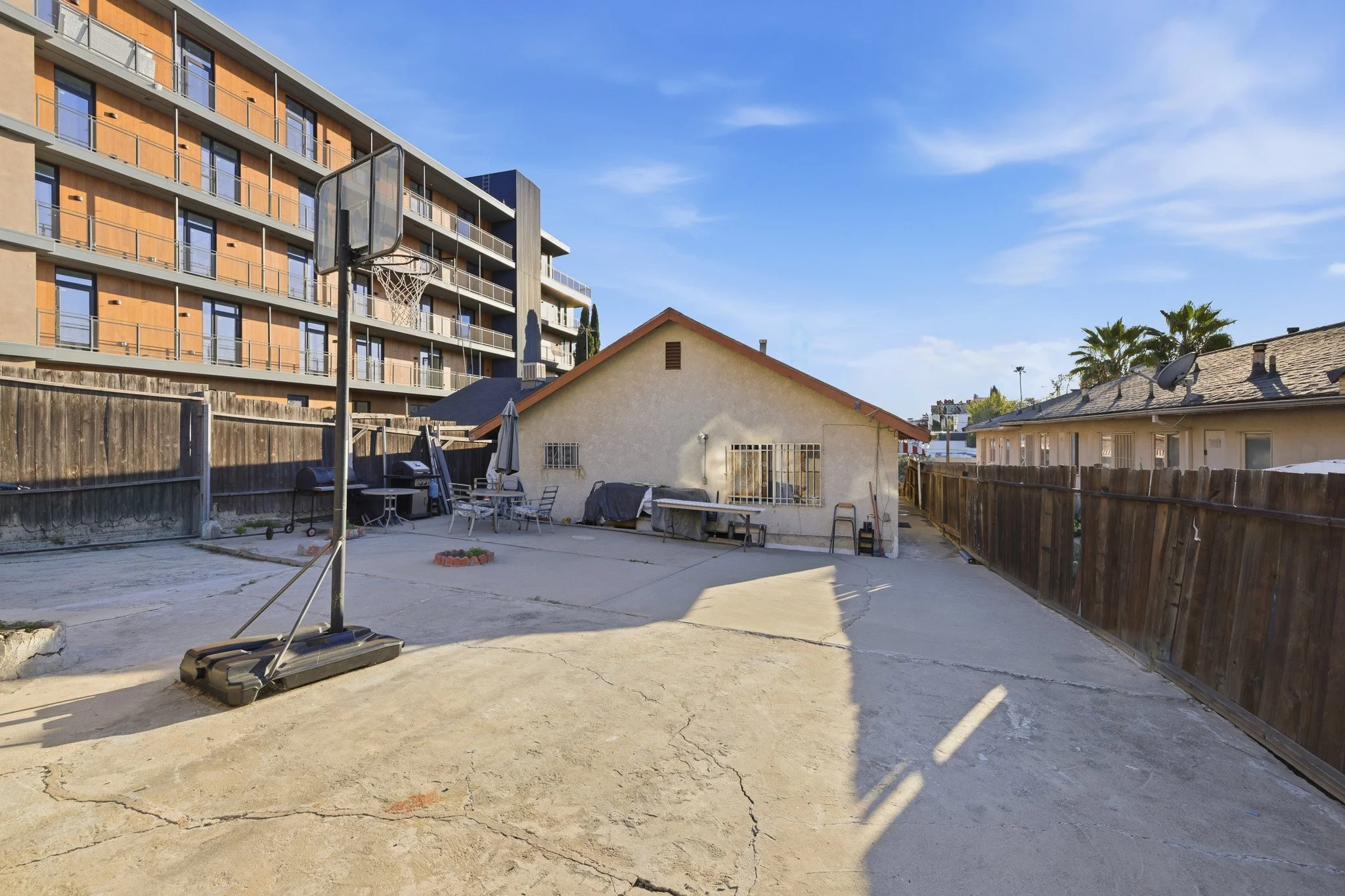 Backyard with concrete pavement, outdoor furniture, a basketball hoop, and a wooden fence. In the background are small houses and a large modern apartment building under a blue sky with some clouds.