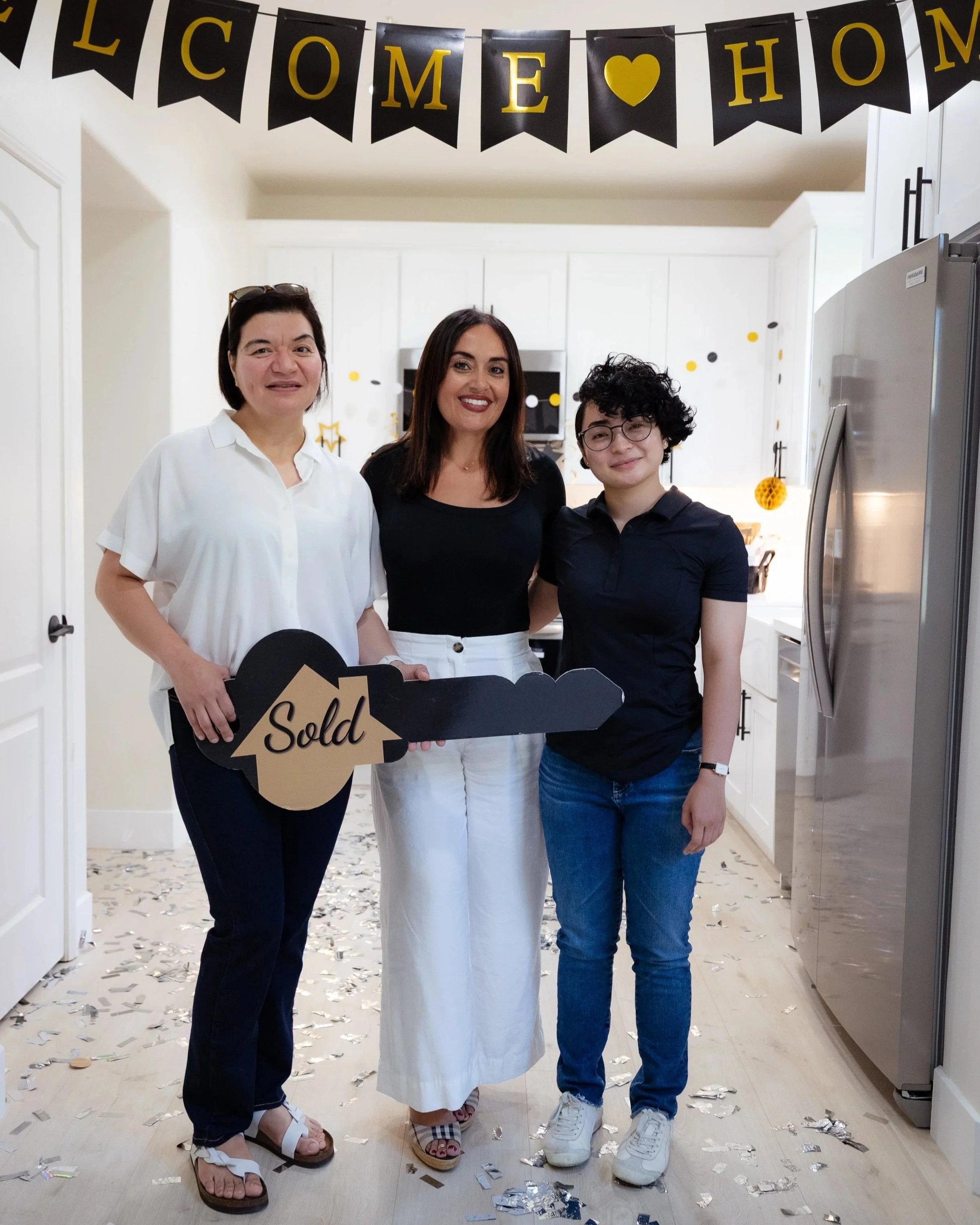 Three women standing together in a kitchen decorated with 'Welcome Home' banner, holding a 'Sold' sign, with confetti on the floor, celebrating a real estate transaction.