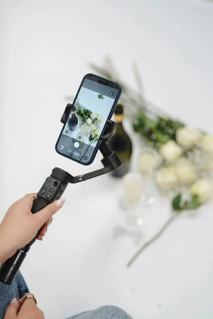 Nahaufnahme einer Hand, die ein Smartphone mit einem Handgimbal hält und ein Blumenarrangement fotografiert, das unscharf im Hintergrund ist.