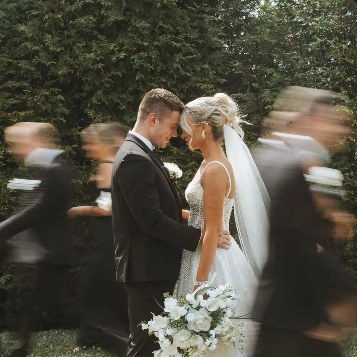 Ein Brautpaar mit ihren Trauzeugen, die sich bei einer Hochzeit umarmen, vor einer grünen Hecke, im Hintergrund sind verschwommene Personen in schwarzen Anzügen.