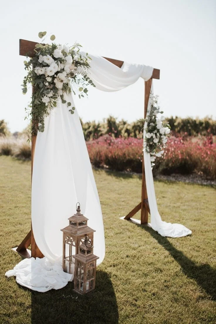 Ein zeremonieller Holzrahmen für eine Hochzeit im Freien, dekoriert mit weißen Tüchern, Blumen und zwei orangefarbene Laternen