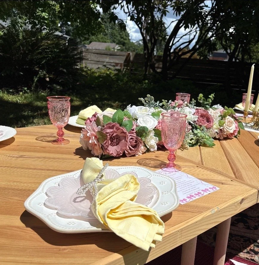 Outdoor tablescape with blush pink floral centerpiece, pink vintage-style goblets, scalloped white plate, and yellow linen napkin tied with pearls, set on a wood table in a garden