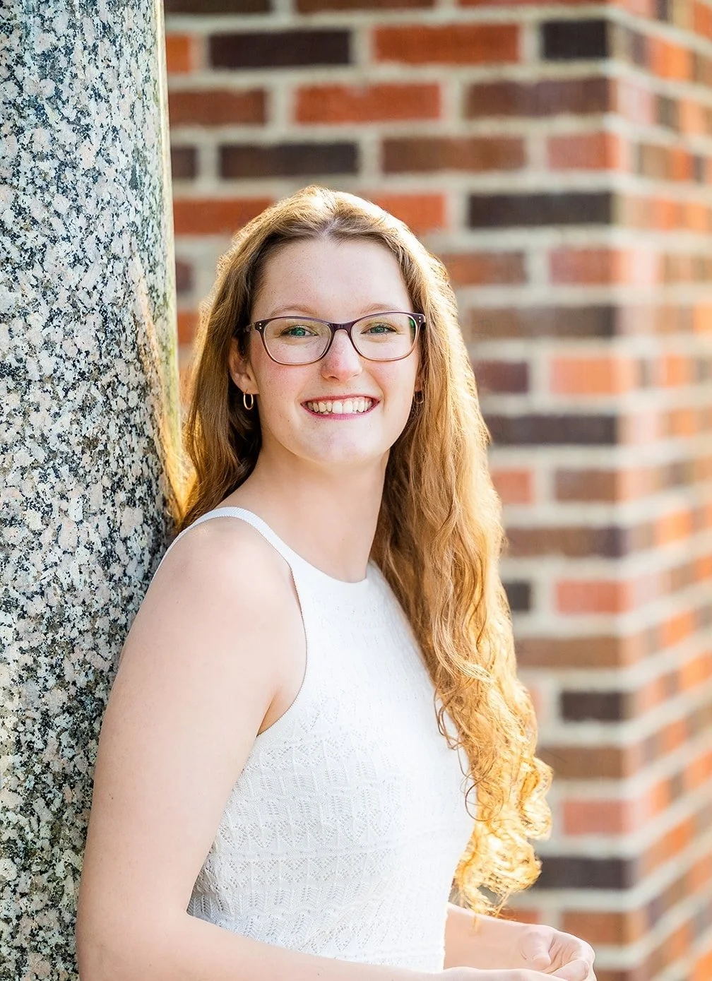 A young woman with long red hair, glasses, and a white sleeveless dress, smiling and standing outdoors next to a textured stone wall and a brick wall in the background.