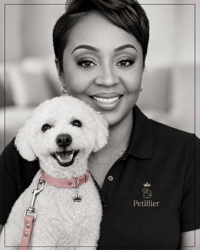 A smiling woman with short hair and makeup, dressed in a black polo shirt with a pet-related logo, poses with a happy white poodle wearing a pink collar and charm, in a professional setting.