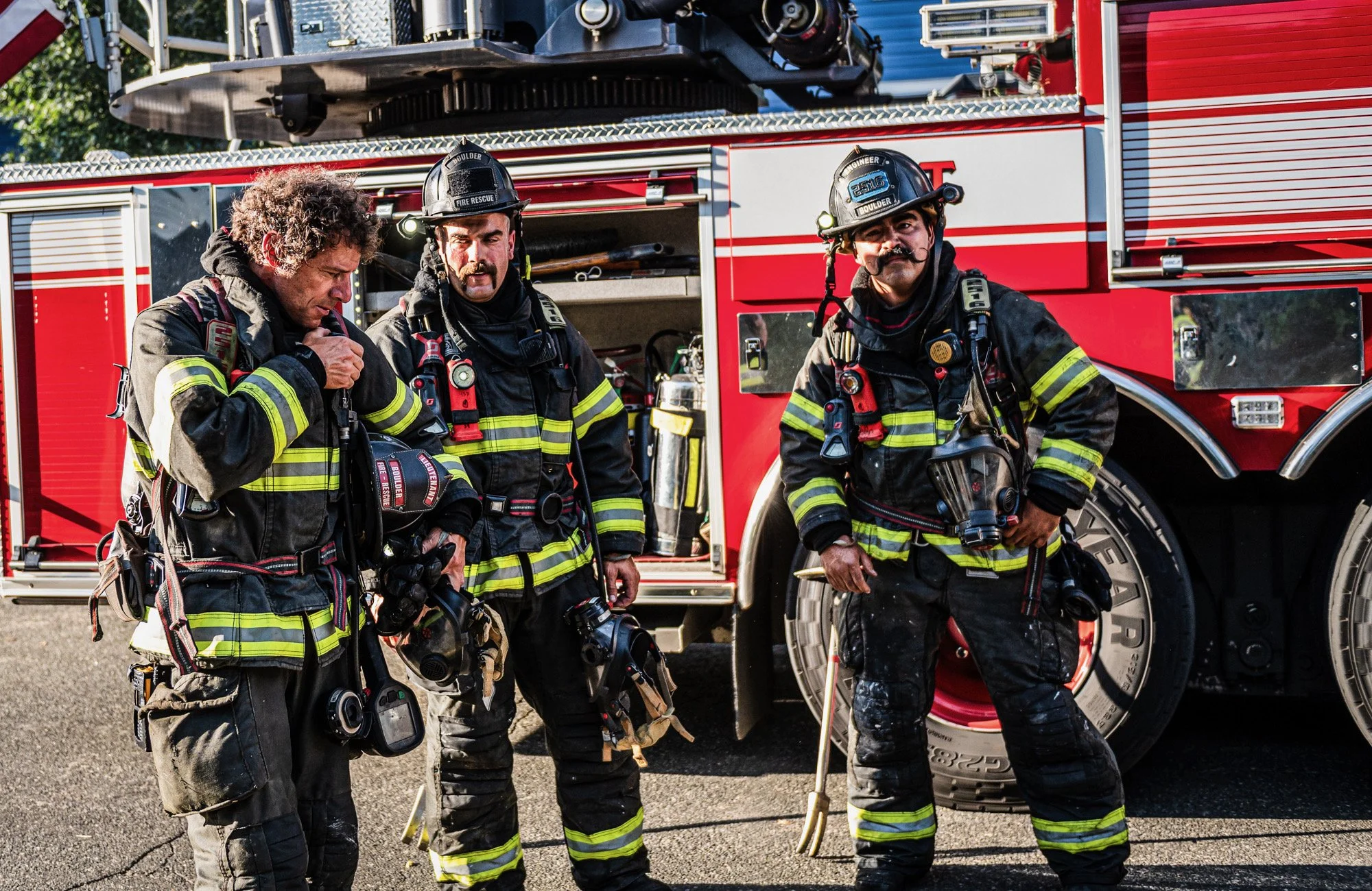 Three firefighters in full gear standing in front of a red fire truck with firefighting equipment.