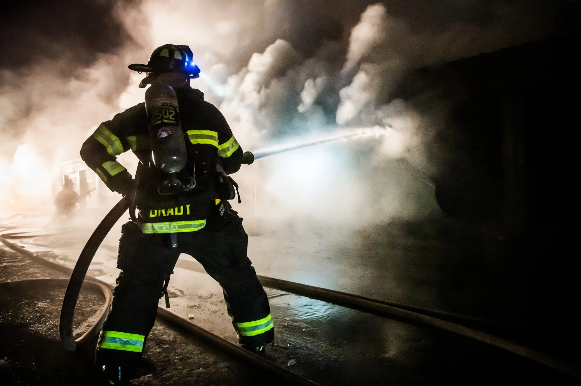 Firefighter in protective gear spraying water at a fire during nighttime, with smoke and flames in the background.
