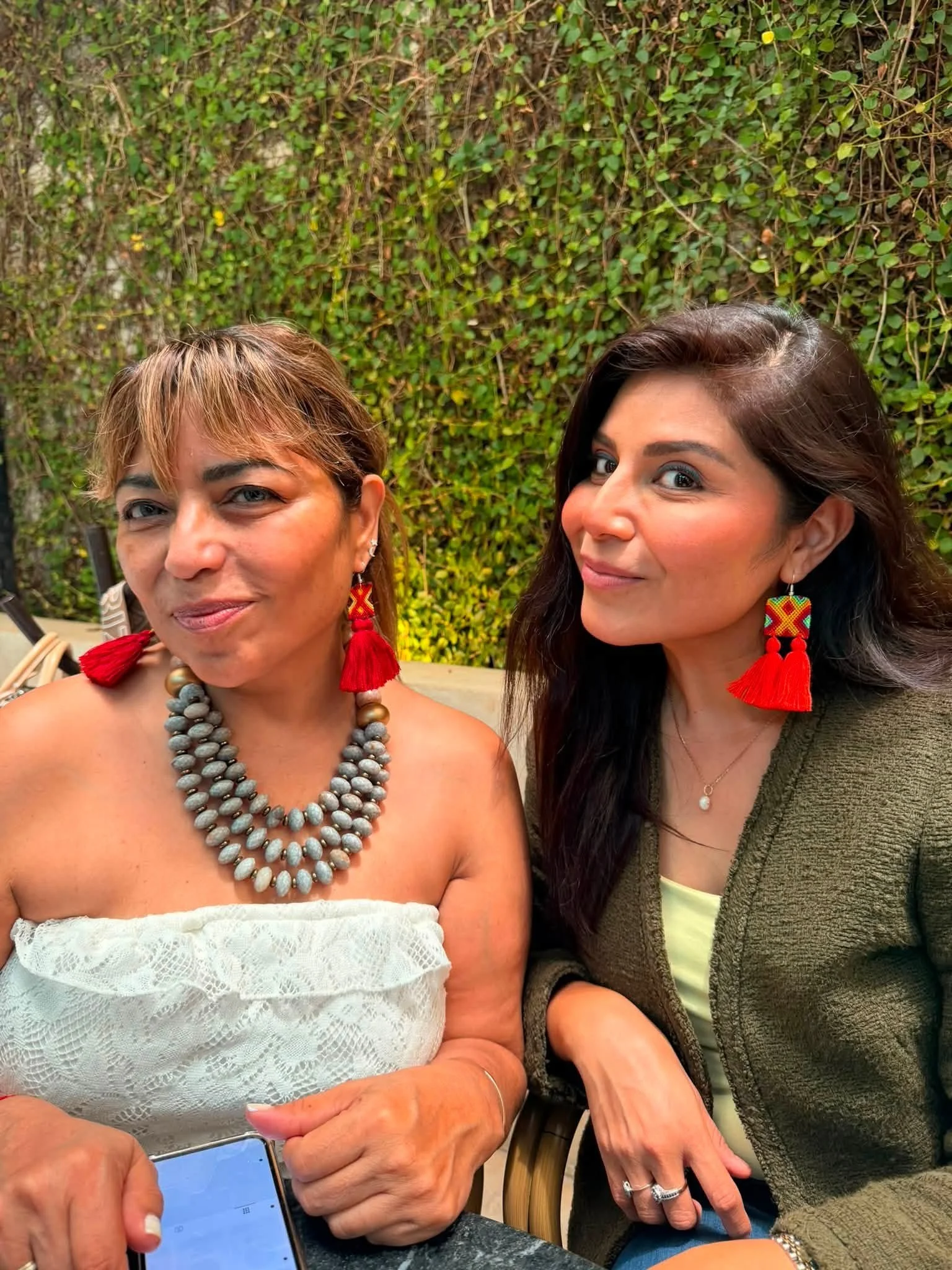 Close-up of two women sitting at a table outdoors with a green leafy background. The woman on the left wears a white lace top, large gray bead necklace, and red earrings, and has short, light brown hair. The woman on the right has long dark hair, wea