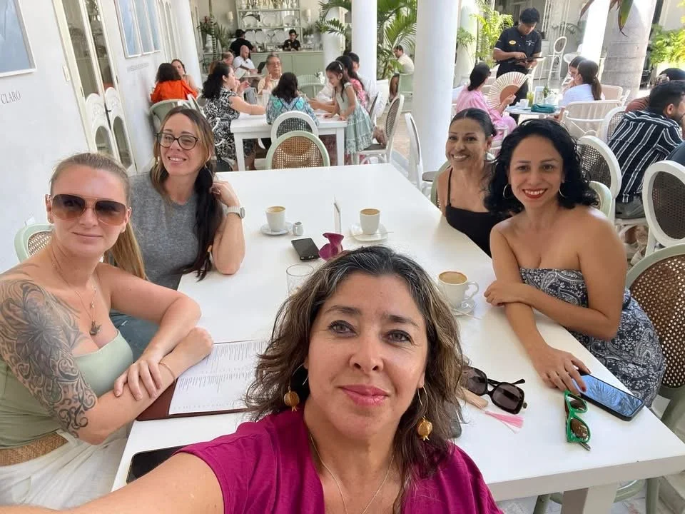 A group of five women sitting at a table in a bright, outdoor cafe, smiling for a selfie. In the background, more people are dining and chatting.