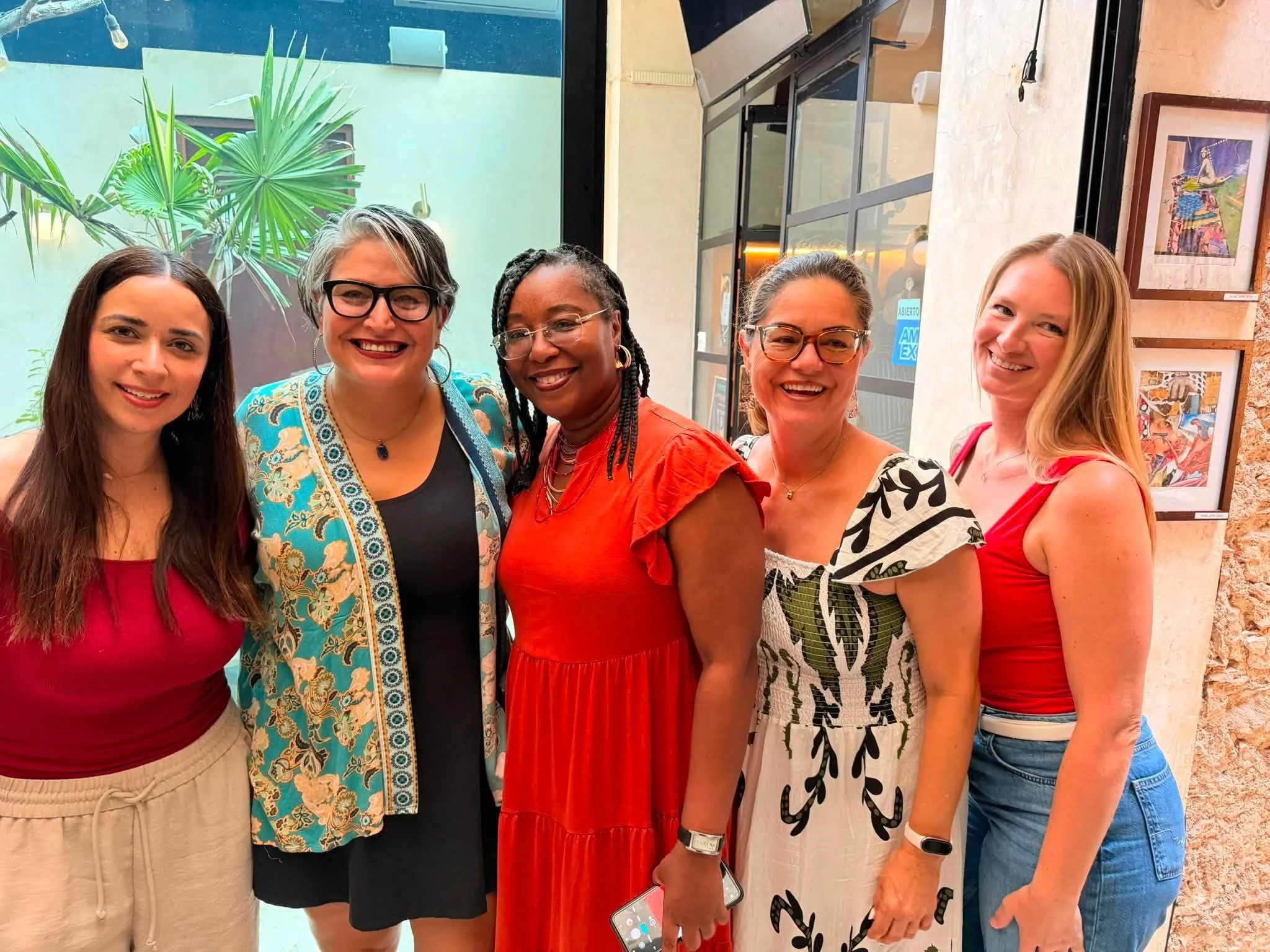 Five women standing side by side indoors, smiling at the camera. The background shows a green wall with a palm plant and an art piece hanging on a brick wall. The women are dressed in colorful, summer-style clothing, and some are wearing glasses.