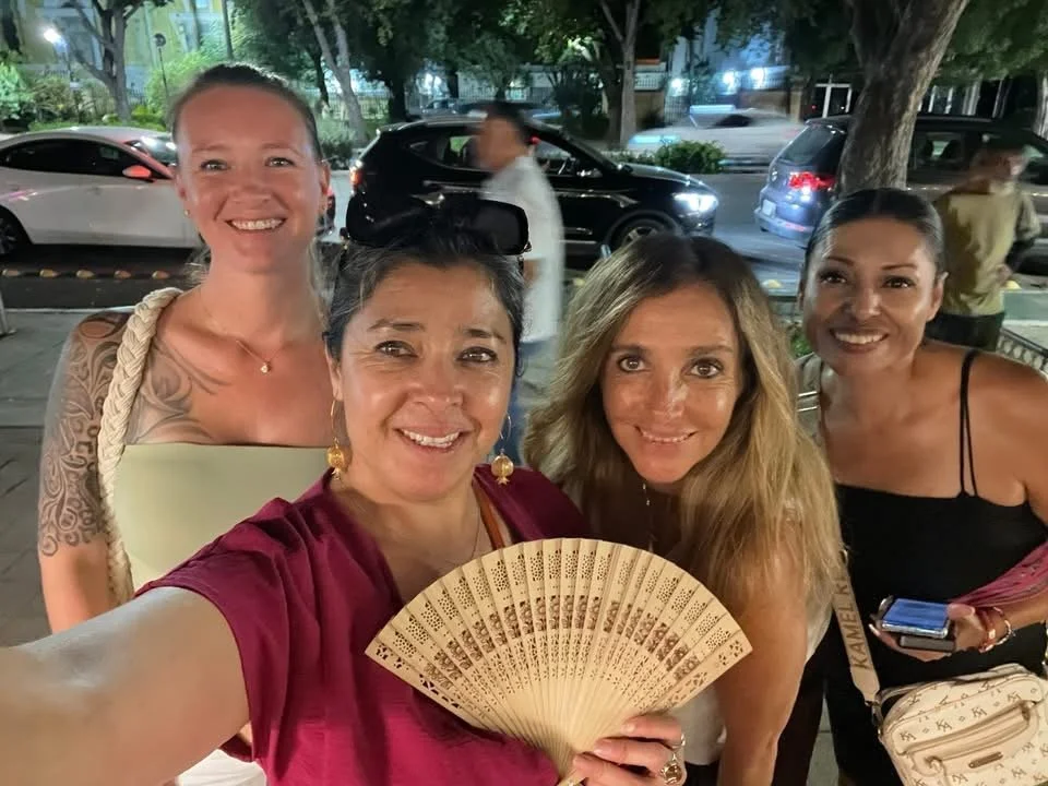 Four women smiling, taking a selfie outdoors at night with parked cars and trees in the background.