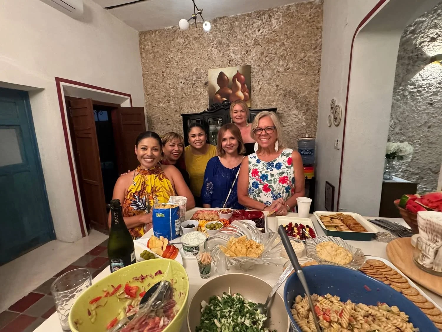 Group of women at a dinner party, smiling around a table filled with various dishes and drinks.