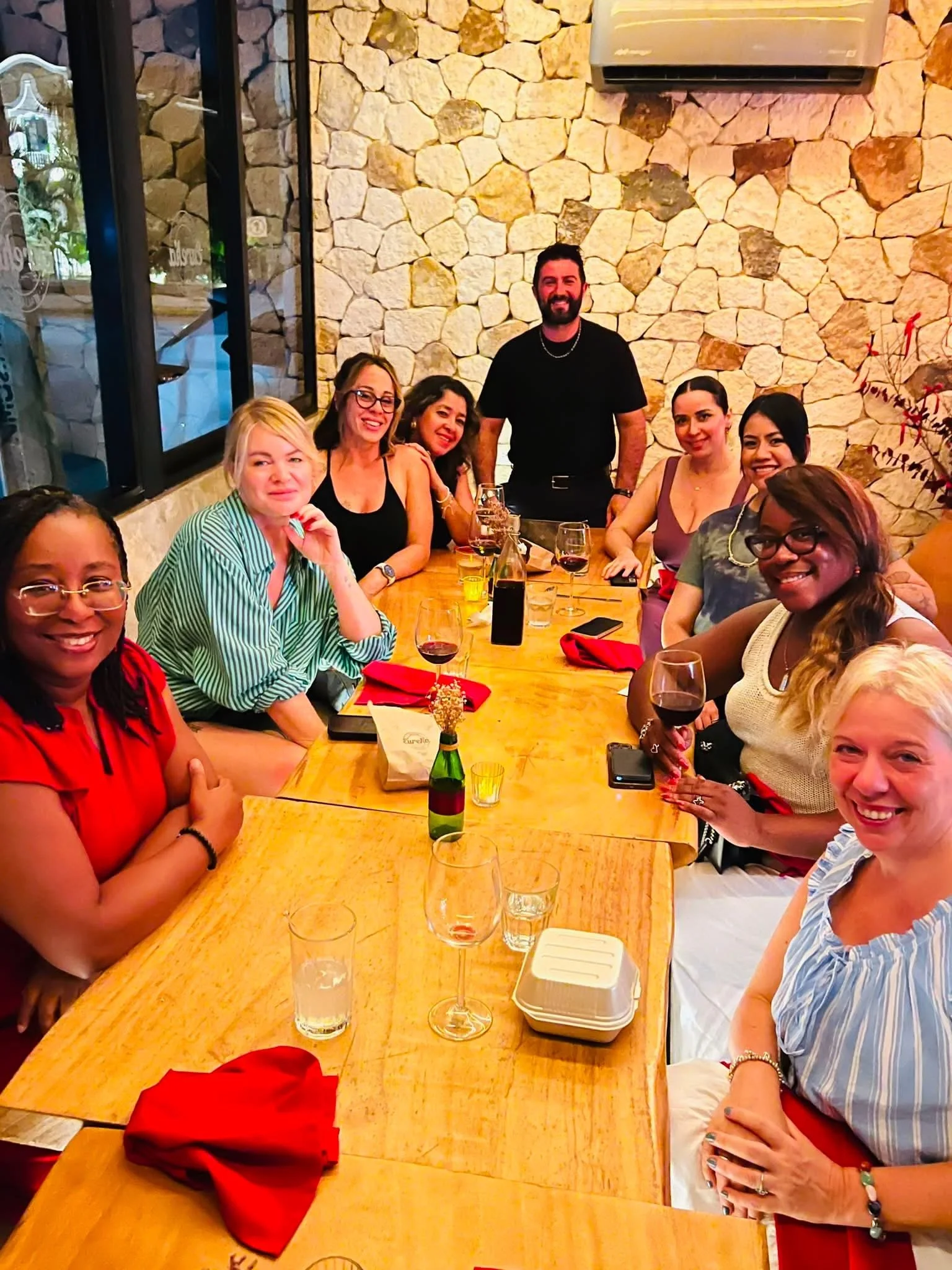 People sitting around a wooden table in a restaurant, smiling and enjoying drinks. The background wall is made of beige and brown stones, and there are red napkins and wine glasses on the table.