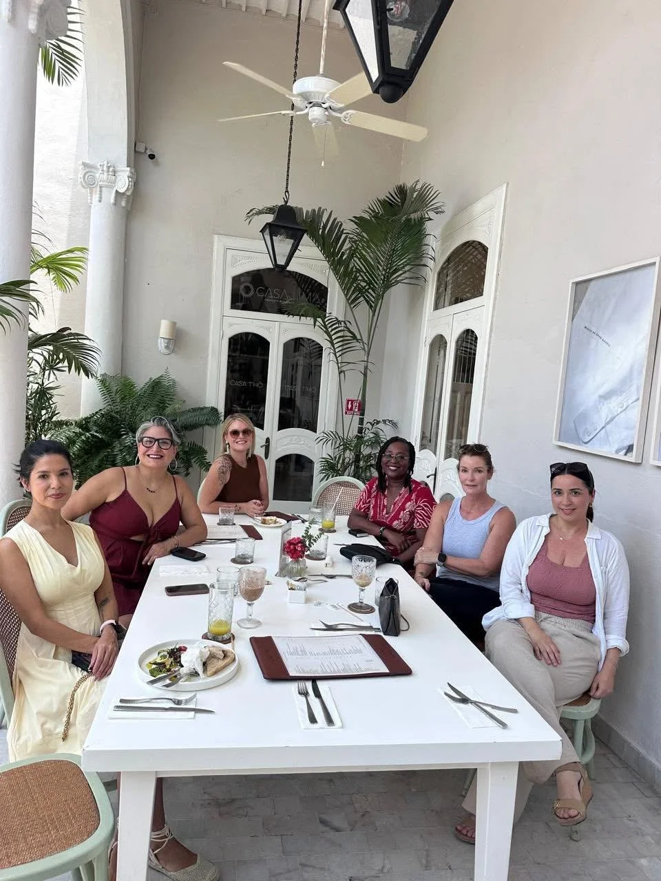 Six women sitting at a white dining table outdoors, with plates, drinks, and menus, surrounded by greenery and white walls.