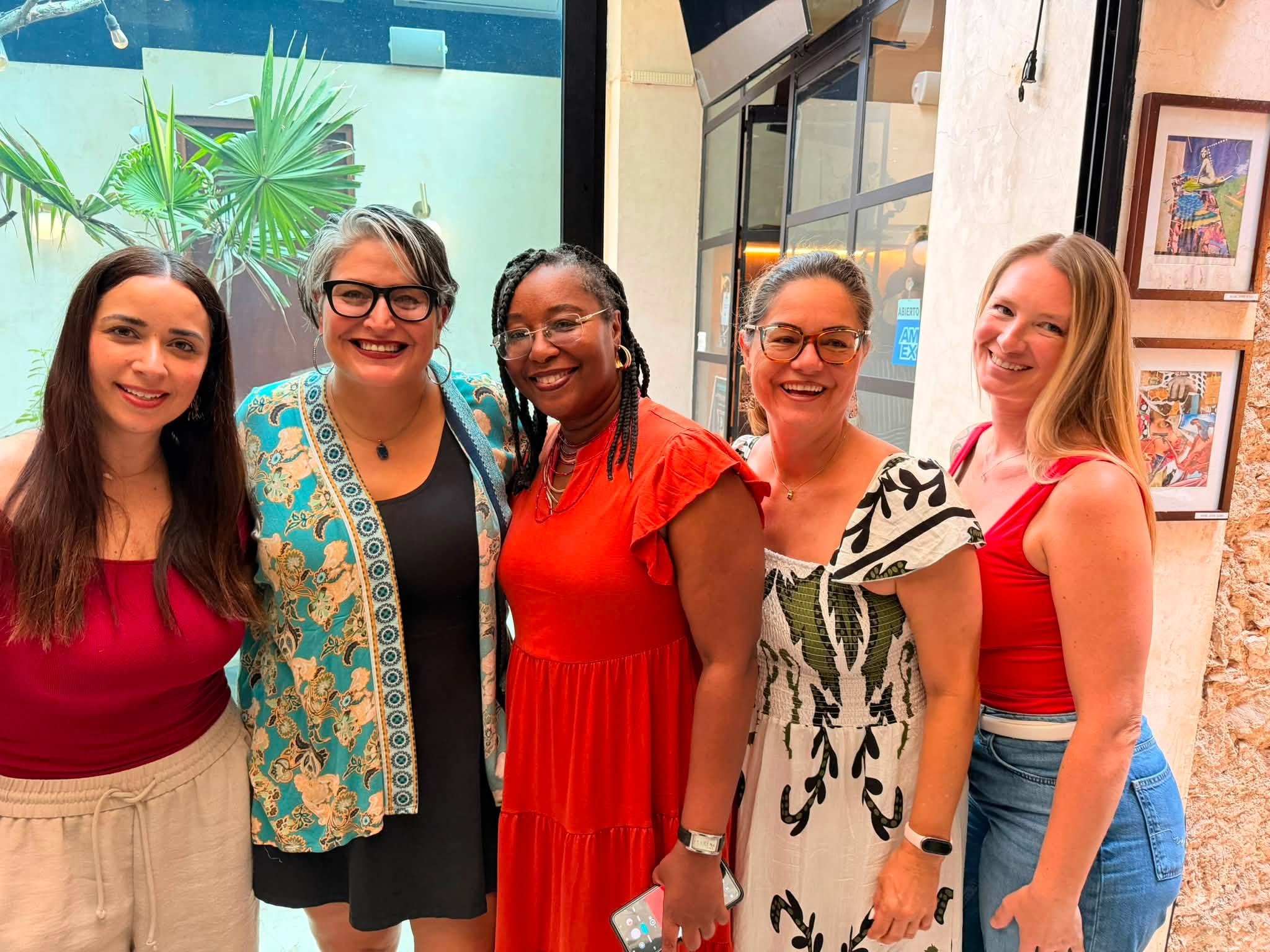 A group of six women standing together indoors, smiling at the camera. The background includes a large plant, framed artwork on the wall, and a glass door with signs.
