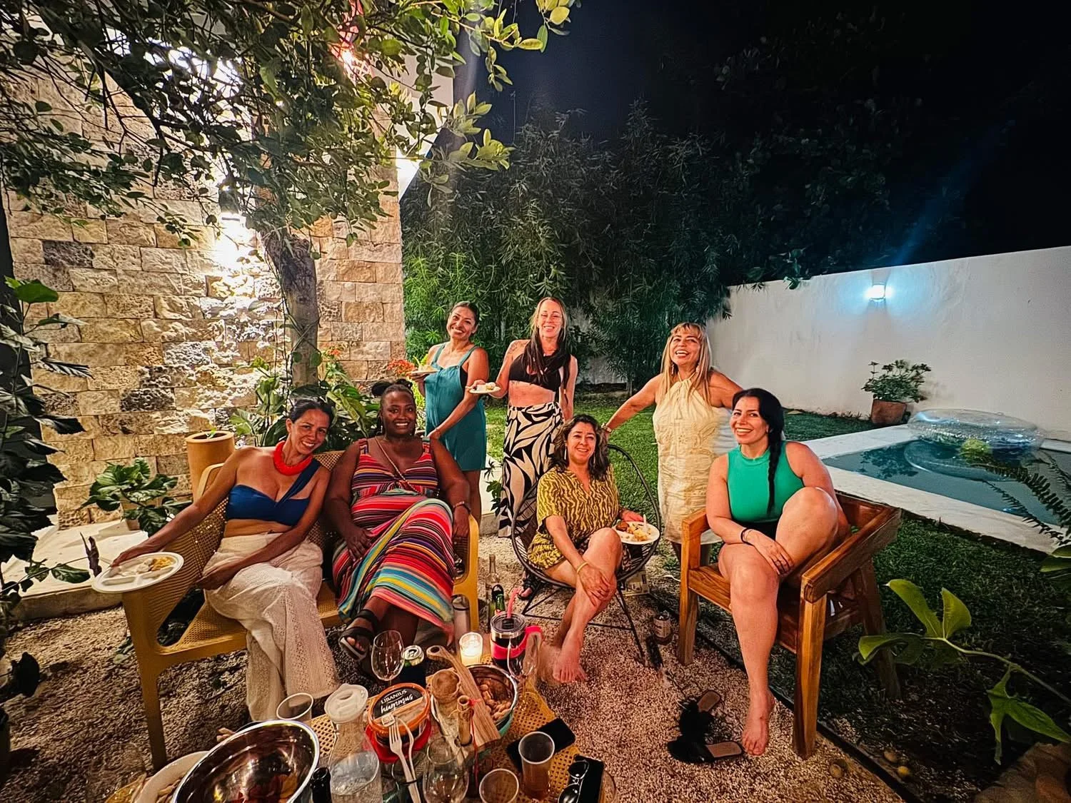 Eight women gathered outdoors at night, smiling and enjoying a social gathering with food and drinks in a garden with a jacuzzi, trees, and lit wall.