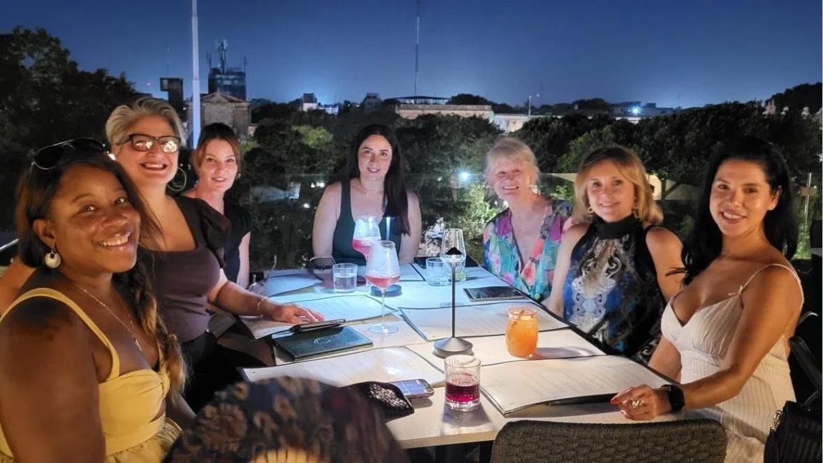 Group of seven women seated at a dinner table outdoors at night, with city skyline and trees in the background, smiling and enjoying drinks.