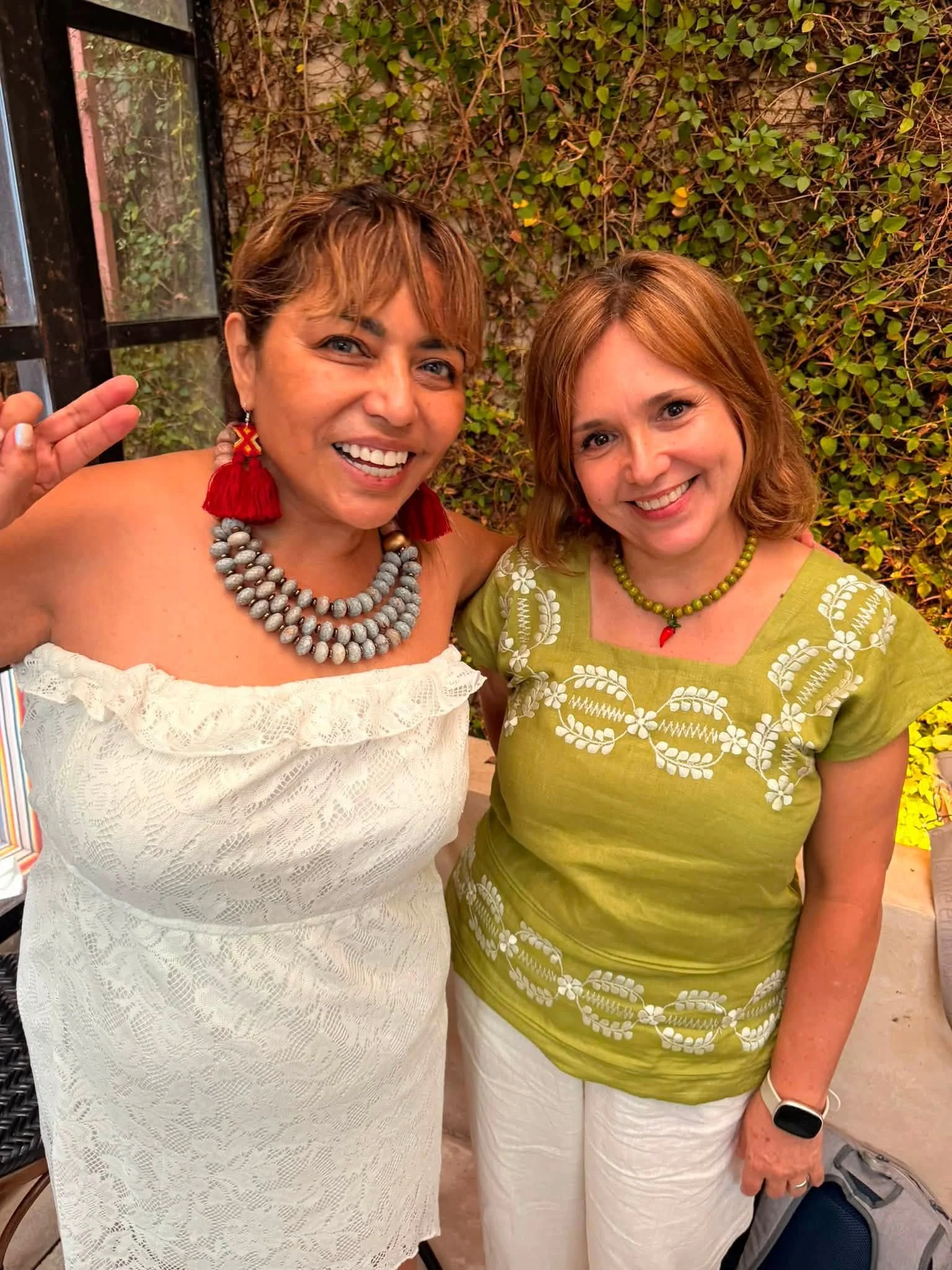Two women smiling and posing for a photo outdoors. One woman has short brown hair, wears a white off-shoulder dress with a beaded necklace and red earrings. The other woman has shoulder-length reddish hair, wears a green embroidered blouse with white