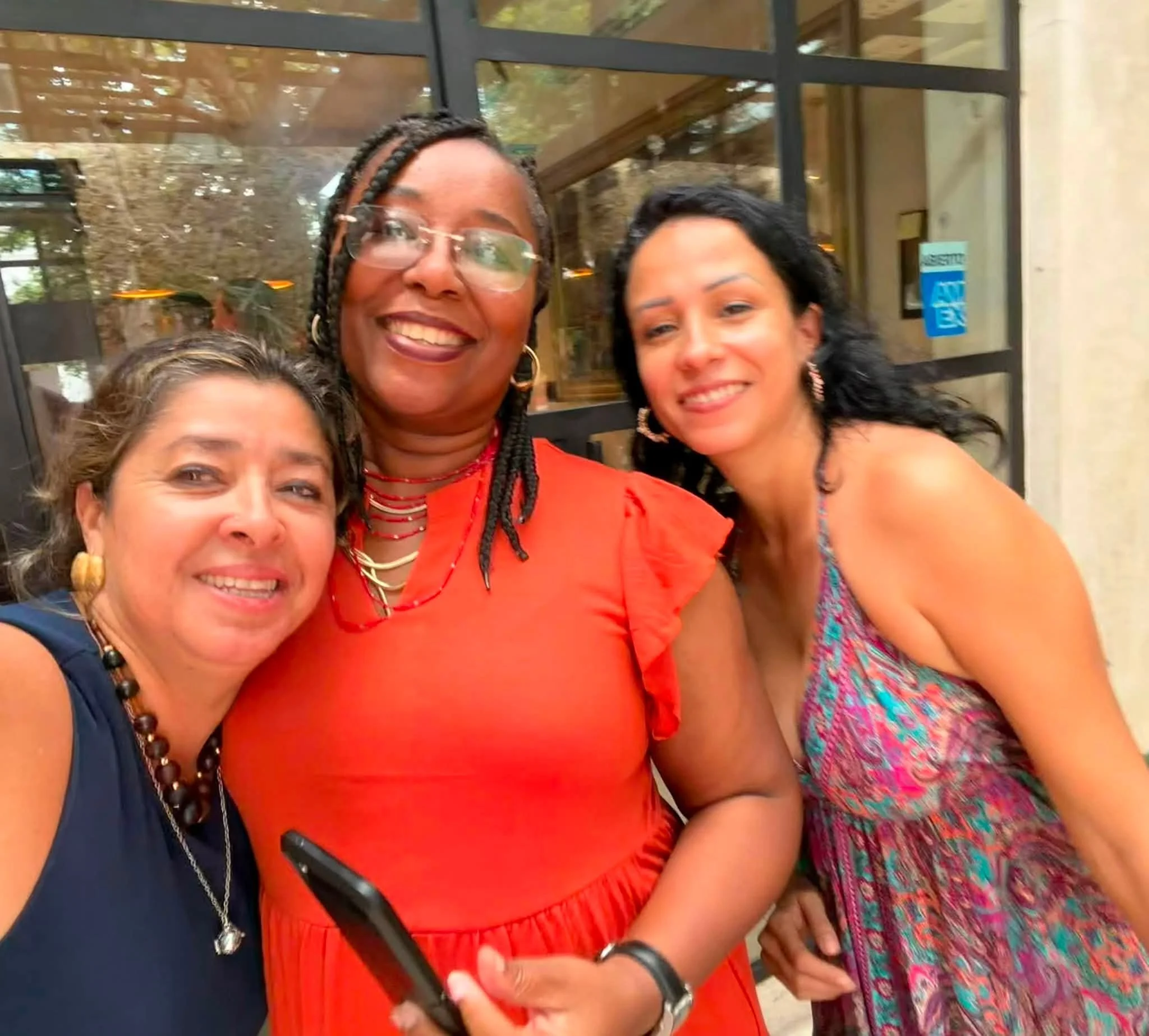 A group of women smiling and taking a selfie, standing in front of a glass door that reflects trees and sky, with a sign marked 'ENTRANCE' visible on the right side of the door.