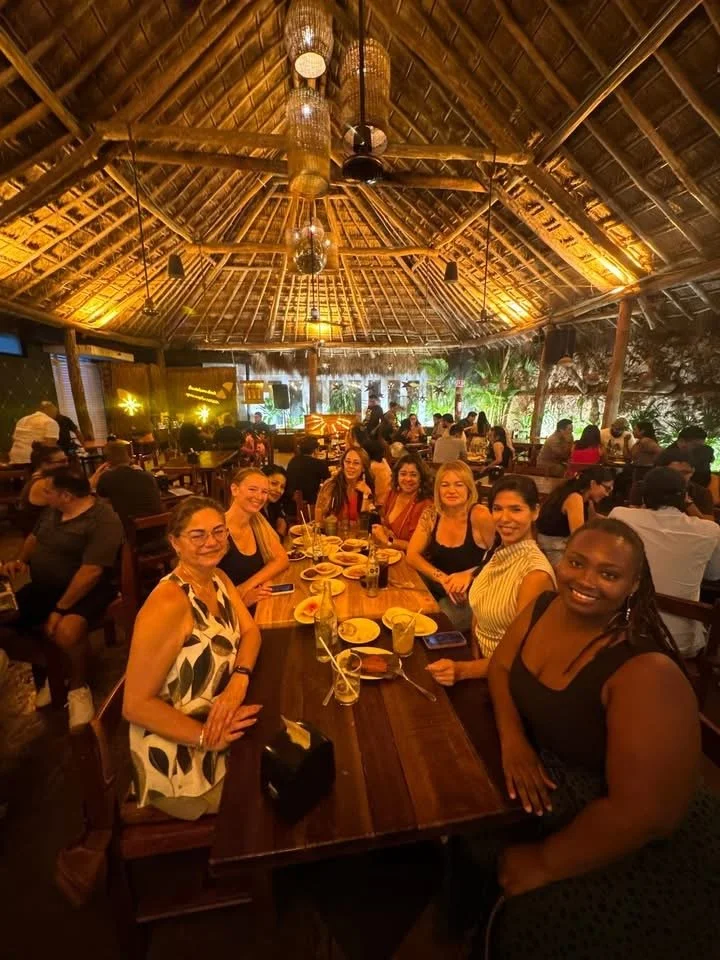 Group of women smiling and sitting around a table with drinks in a tropical-style restaurant with a thatched roof and warm lighting.