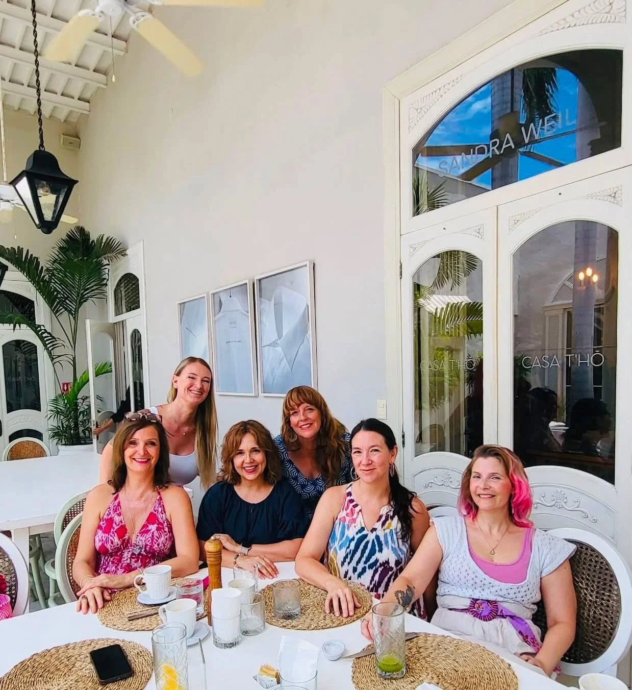 Six women sitting and standing around a table in a restaurant, smiling. The table has drinks, cups, and placemats. The restaurant has large white doors, potted plants, and modern artwork.