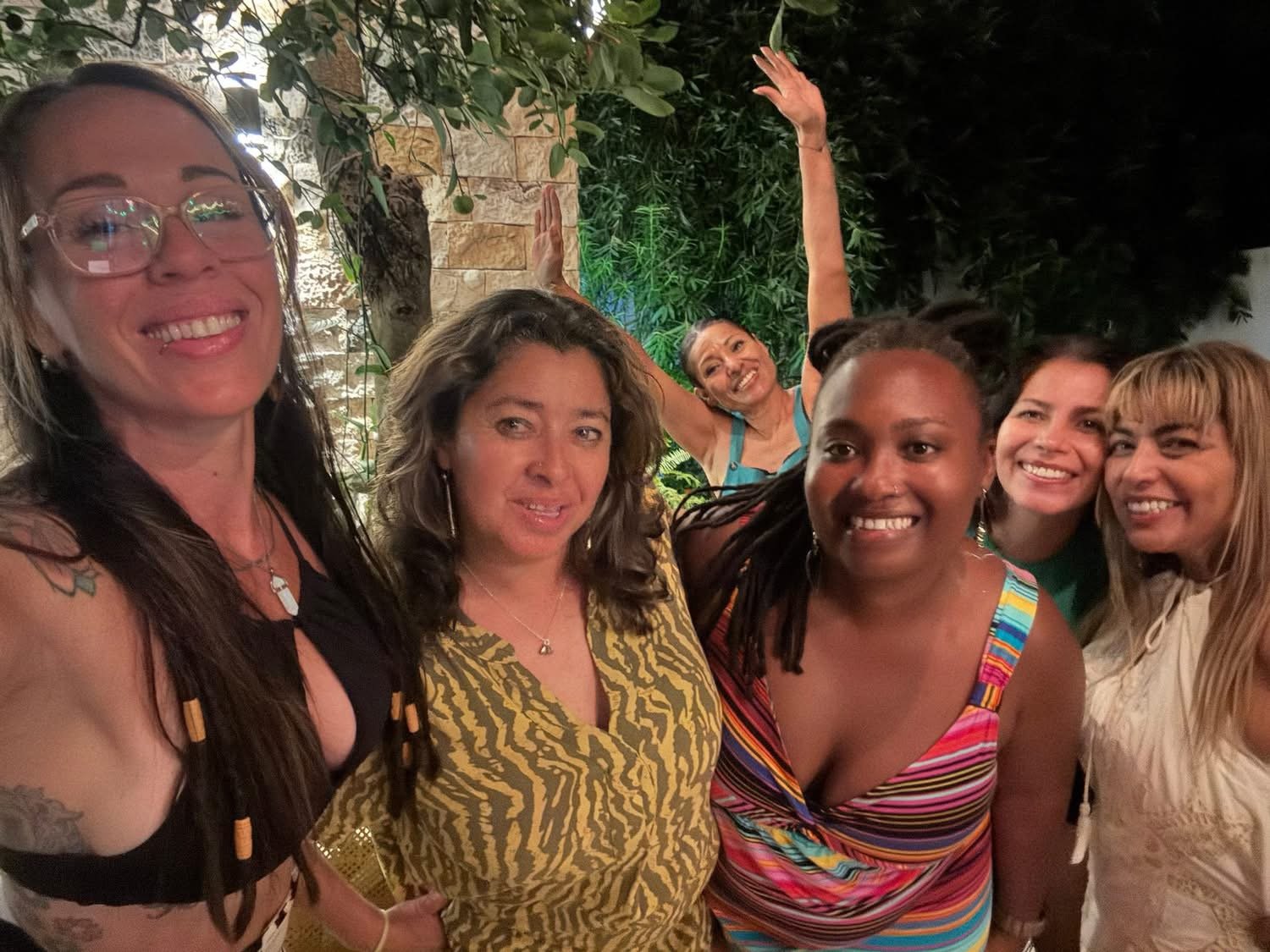 Group of six women smiling and posing at an outdoor gathering at night, surrounded by greenery.