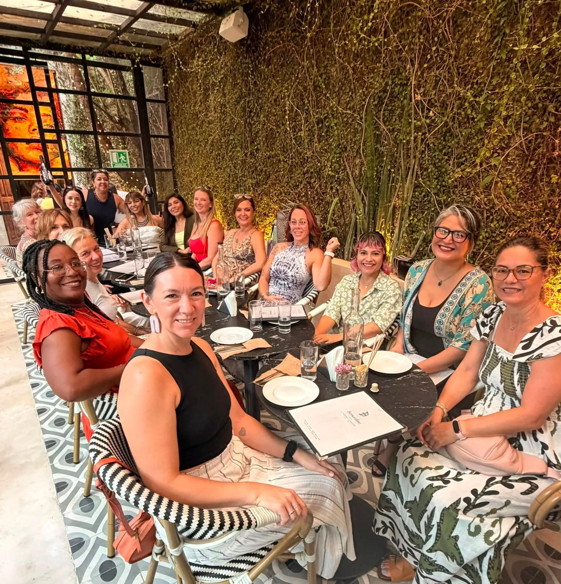 Group of women sitting at a long table in a restaurant, smiling and enjoying a meal. The setting includes patterned chairs, glasses, plates, and a large green indoor wall with plants. Some women are wearing colorful dresses, and there are glasses and plates on the table.
