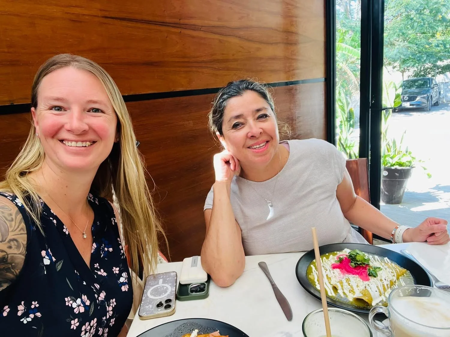 Two women sitting at a restaurant table smiling for the camera, with food and drinks on the table, and a large window showing greenery and parked cars outside.